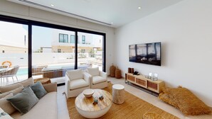 A modern living room with a large window, a flat-screen TV, a sofa, a coffee table, and a wicker console. - Kalliopi is a contemporary villa, at Konnos Beach , within Periyiali Resort. (Protaras)