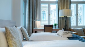 Une chambre d’hôtel avec un lit, un bureau et une chaise. Il y a une fenêtre avec des rideaux et une lampe sur le bureau.