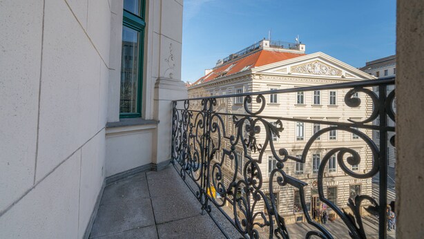 Een balkon met gedetailleerde, zwarte ijzeren hekwerken, een wit gebouw met groene raamkozijnen, en een gebouw met een rood dak op de achtergrond.