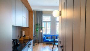 Et moderne køkken med en sort bordplade, hvide skabe og et træloft. Et spiseområde med en blå sofa og et lille bord er synligt gennem en åben dør.