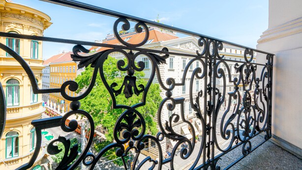 En balkon med detaljerede, sorte metalgelændere med udsigt over en gade med historiske bygninger og et træ.