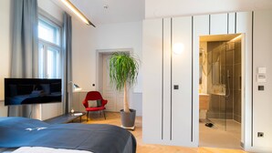A modern hotel room with a bed, TV, red chair, potted plant, and a glass-enclosed shower area.