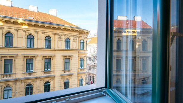 A view through a window of a building with classical architecture, featuring multiple windows and a red-tiled roof.