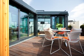 En moderne udendørs terrasse med glasdøre, trægulv og hvide fletmøbler.