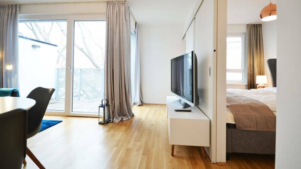 A modern hotel room with a large window, a flat-screen TV on a white stand, a bed with a grey headboard, and a dining area with a table and chairs. - Vienna Residence, Donaust.- United Nations #6251 (Vienna)