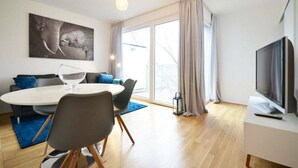 A modern living room with a round dining table, grey sofas, a flat-screen TV, and a large window with curtains. - Vienna Residence, Donaust.- United Nations #6251 (Vienna)