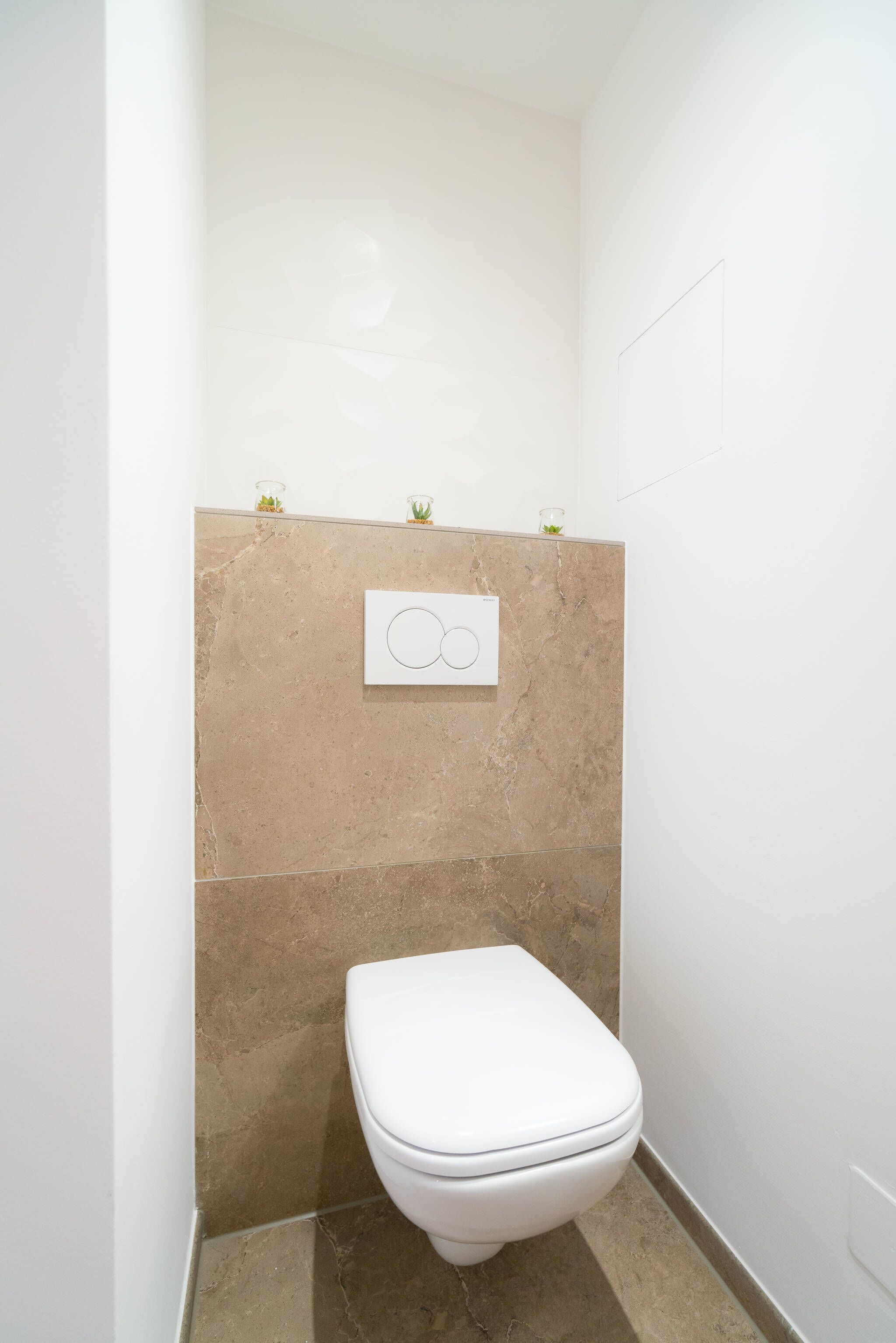 Een compacte, moderne badkamer met een wit toilet, beige tegels en een bedieningspaneel voor de spoeling aan de muur.