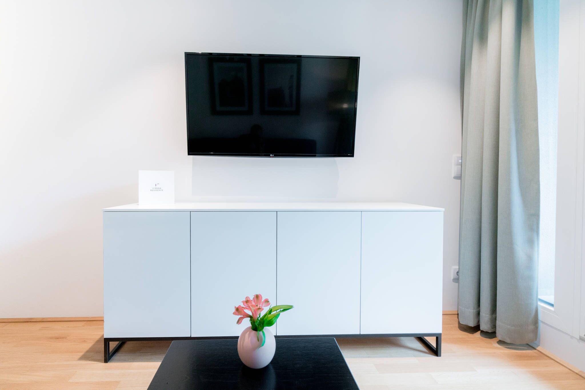 Un soggiorno moderno con una TV a schermo piatto montata a parete, un mobile bianco, un tavolino da caffè nero e un vaso con fiori.