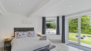 A bedroom with a large bed, a nightstand, a bedside table, a window with curtains, and a view of the outdoors. - Stunning, Contemporary 1 Bedroom En-suite Annexe (Plymouth)