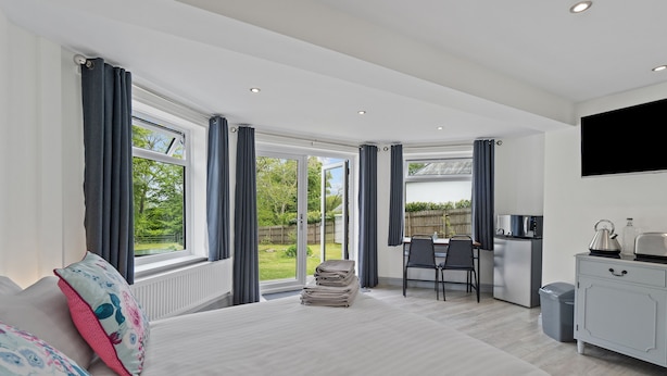 A modern bedroom with a large bed, a desk, and a view of the garden through the windows. - Stunning, Contemporary 1 Bedroom En-suite Annexe (Plymouth)