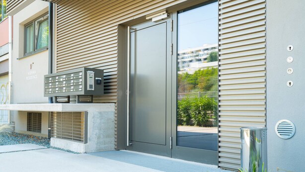 A modern building with a metal door, a bench with a mailbox, and a small garden area with pebbles and a plant. - Vienna Residence, Hietzing - Schönbrunn #6375 (Vienna)