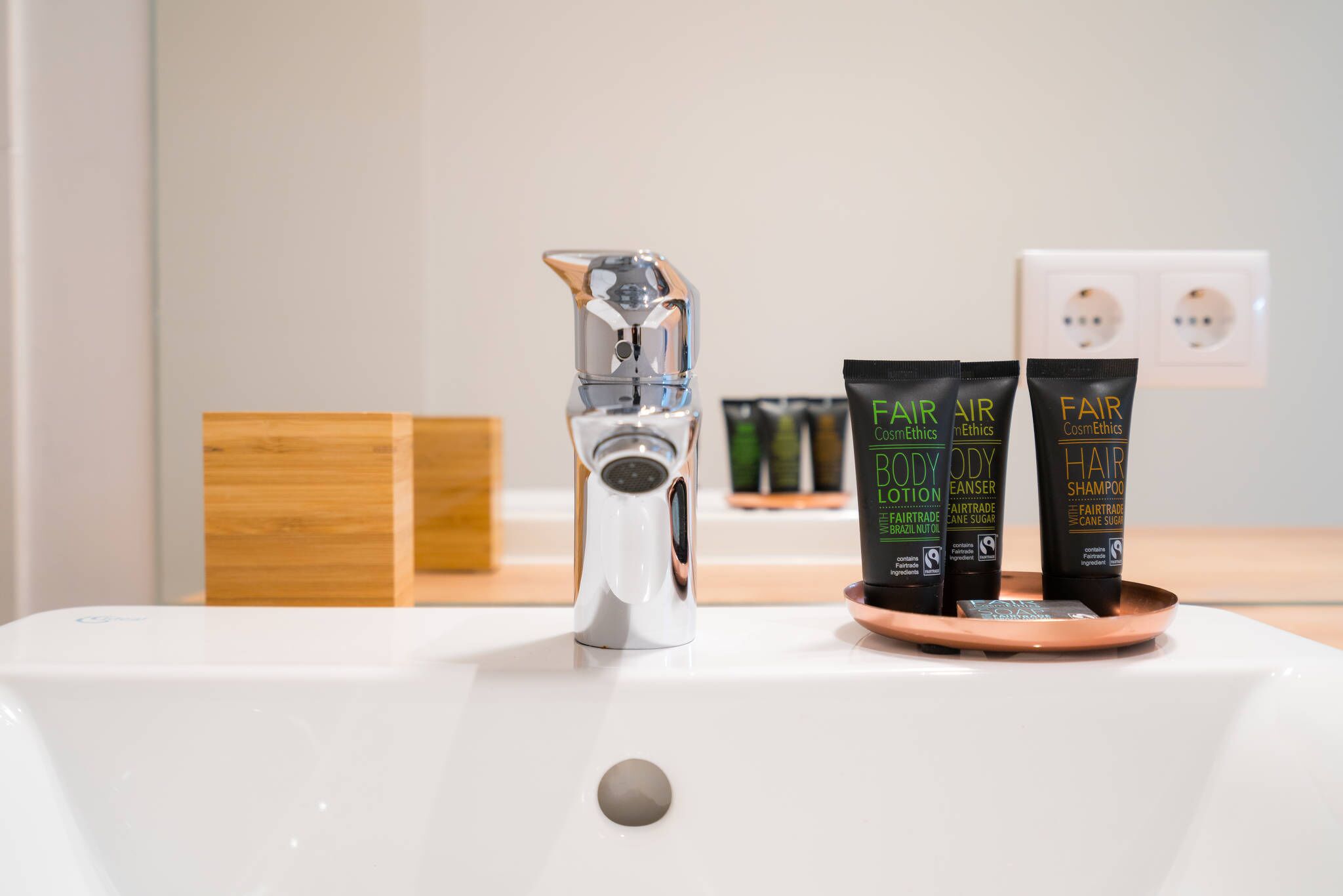 Ein Badezimmerwaschbecken mit einem modernen Wasserhahn, drei Fair Trade Kosmetikprodukte und einer hölzernen Seifenschale.