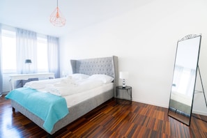 Een netjes opgemaakt bed met een grijze hoofdbord, een nachtkastje met een lamp, een grote spiegel en een raam met dunne gordijnen.