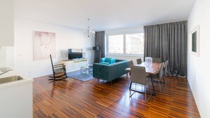 En moderne stue med sofa, spisebord og stole. Der er et fjernsyn og et lille tekøkken med vask.