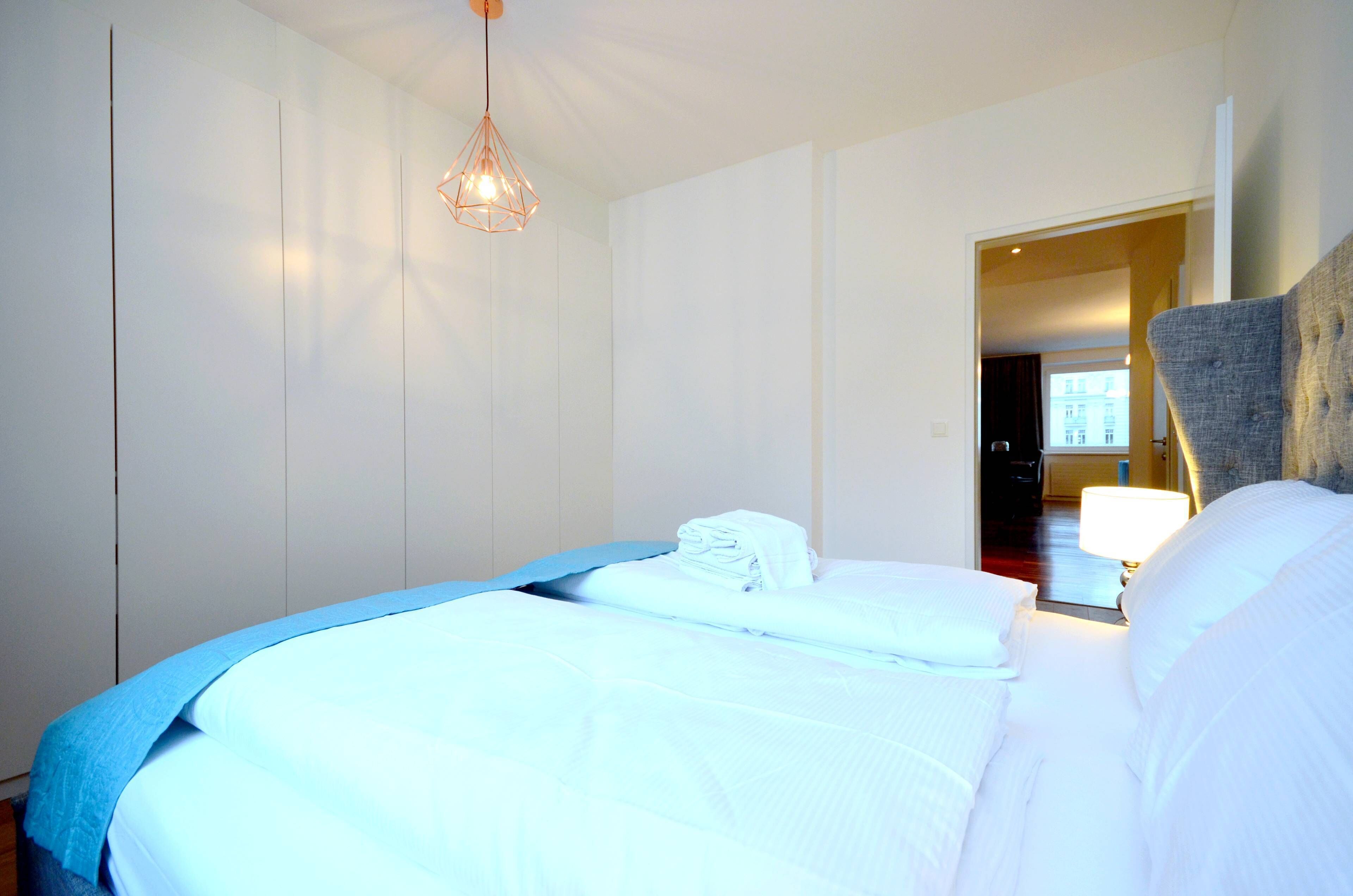 Quarto de hotel com uma cama bem arrumada, um lustre pendurado e vista para o corredor.