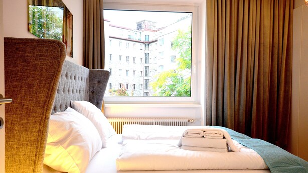 Et hotelværelse med seng, gardiner foran et vindue, en radiator og udsigt til en bygning.