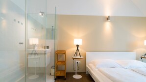 A modern hotel room with a glass shower enclosure, a bedside table with a lamp, a wooden shelf, and a neatly made bed. - Vienna Residence, City Park - Belvedere #2535 (Vienna)