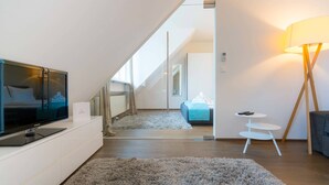 A modern, well-lit room with a flat-screen TV on a white cabinet, a fluffy gray rug, a small white side table, and a lamp with a wooden base. - Vienna Residence, City Park - Belvedere #2534 (Vienna)