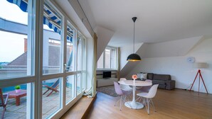 A modern living space with large windows, a dining area with a round table and chairs, a gray sofa, and a black pendant light.
