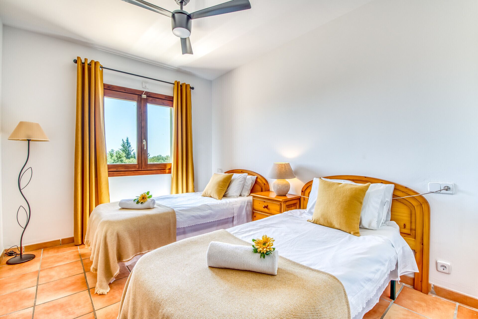 A hotel room with two beds, a ceiling fan, a window with curtains, a bedside table with a lamp, and a floor lamp.