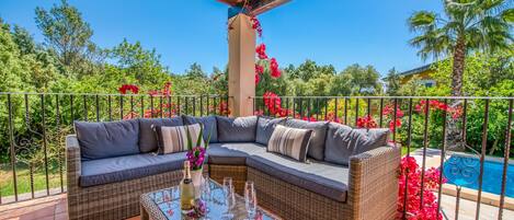 A covered outdoor seating area with a wicker sofa, a glass-top coffee table, and a view of a swimming pool.