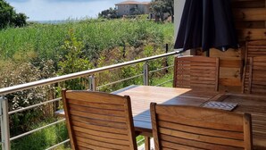 Un patio al aire libre con mesa y sillas de madera, baranda de metal y vistas a un campo y edificios a la distancia.