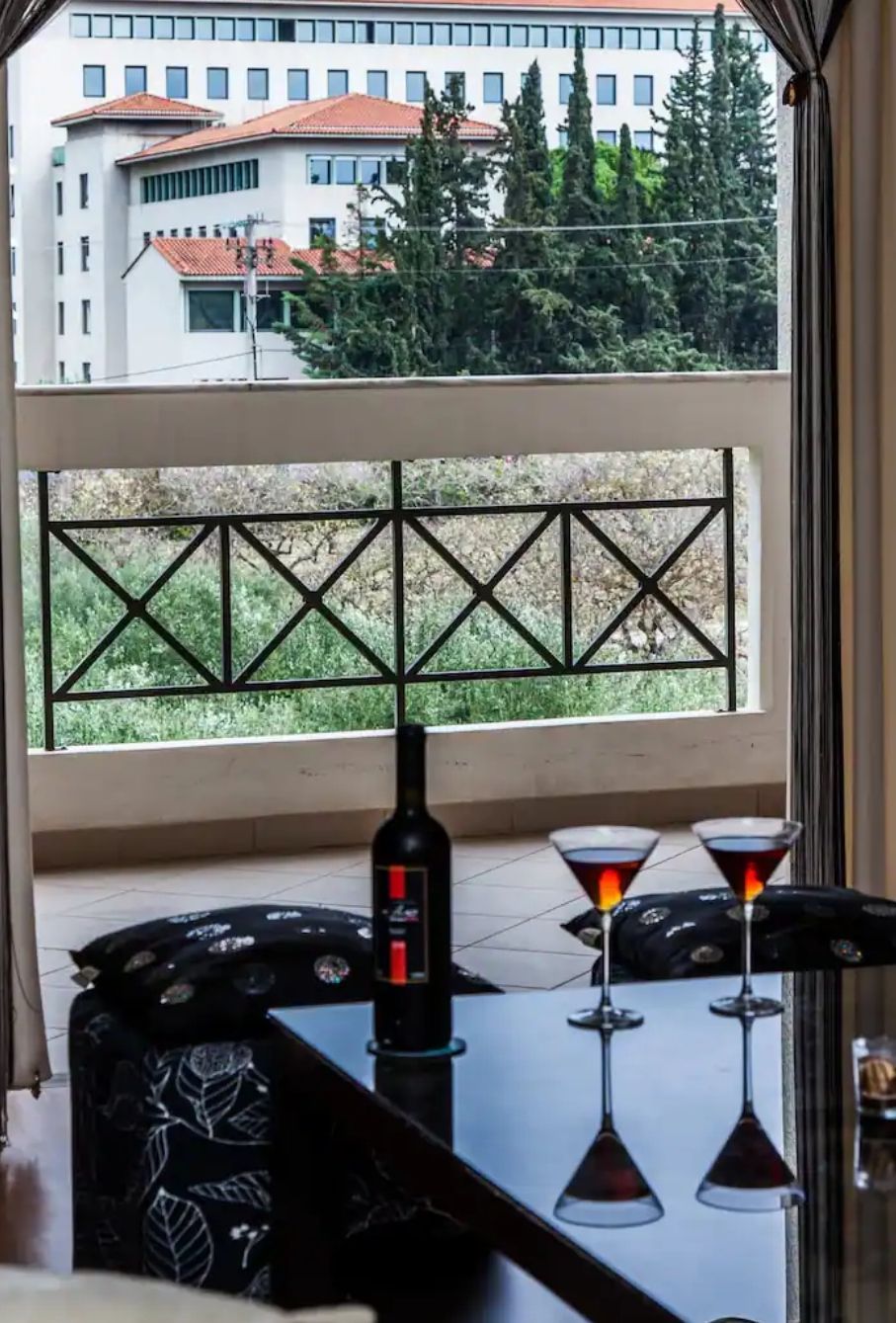 A table with a bottle and two glasses, a window with a view of buildings and trees, and a geometric metal railing.