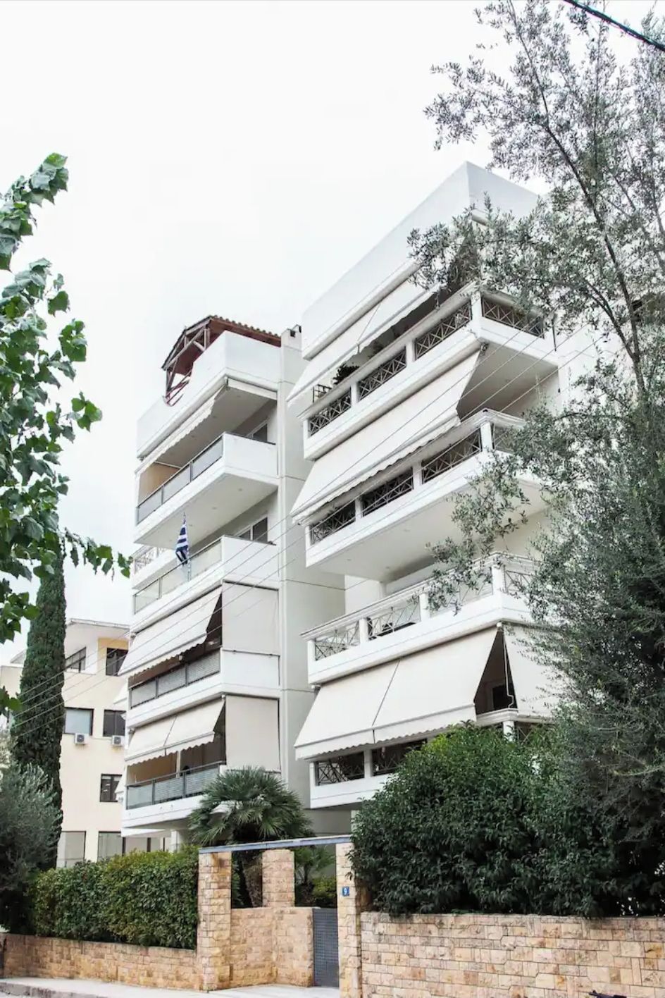 A multi-story residential building with balconies, a stone wall, and surrounding greenery.