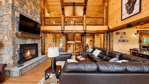 A log cabin interior with a stone fireplace, leather seating, and a staircase leading to an upper level. - Grand Peaks Lodge, Luxury Log Home, Views, Hot Tub, Sauna, EV Charger, 3D Tour (Jay)
