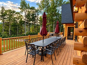 A wooden deck with a long table and chairs, surrounded by a wooden railing. - Grand Peaks Lodge, Luxury Log Home, Views, Hot Tub, Sauna, EV Charger, 3D Tour (Jay)