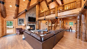 A spacious living room with a stone fireplace, leather sofa, wooden flooring, and a wooden staircase. - Grand Peaks Lodge, Luxury Log Home, Views, Hot Tub, Sauna, EV Charger, 3D Tour (Jay)