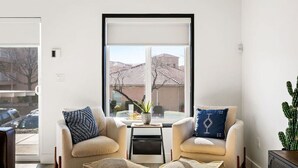 Un salon moderne avec deux fauteuils, une table basse et une grande fenêtre donnant sur des immeubles et des arbres.