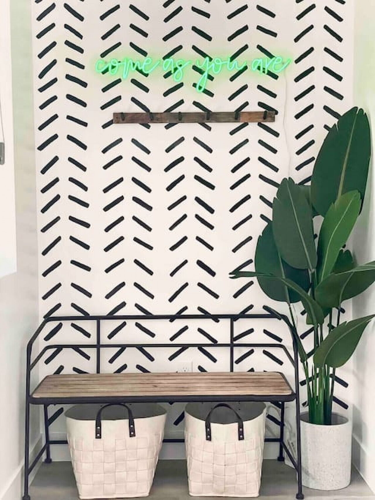 A bench with two storage baskets underneath, a potted plant to the right, and a wall with a diamond pattern and a neon sign above.