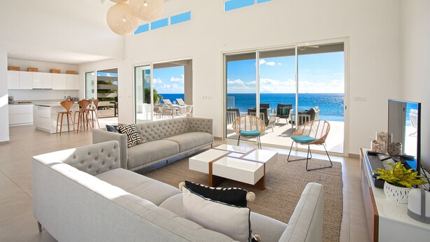 A spacious living area with a large sofa, a coffee table, and a TV. There is a balcony with a view of the ocean. - Aqua (4 bedrooms) - Ocean view villa with pool, Indigo Bay, St. Maarten (Indigo Bay)