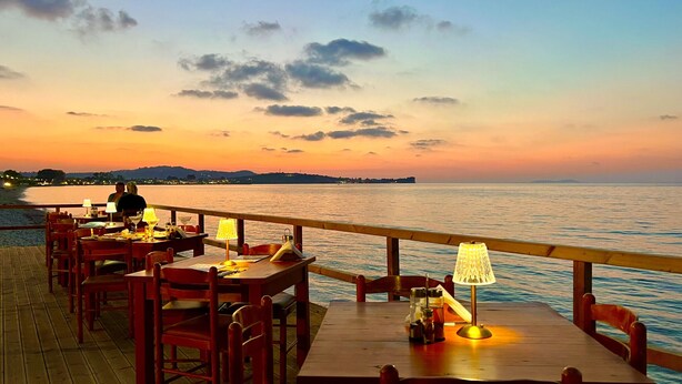 Un restaurant en bord de mer, avec des tables et des chaises, une terrasse en bois et une vue sur la mer et les collines lointaines, particulièrement belle au coucher du soleil.