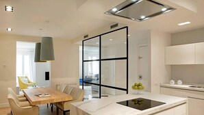 A modern kitchen with a central island, a dining area with a wooden table and chairs, and a view through a glass partition to a living space.