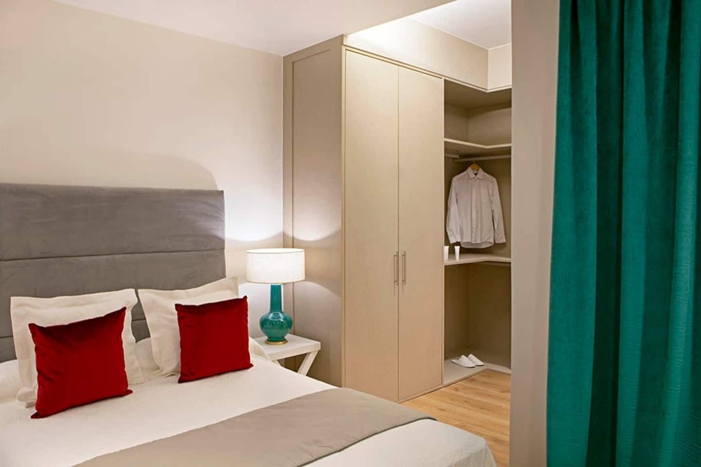 Habitación de hotel con cama, mesita de noche con lámpara, armario y cortina verde.