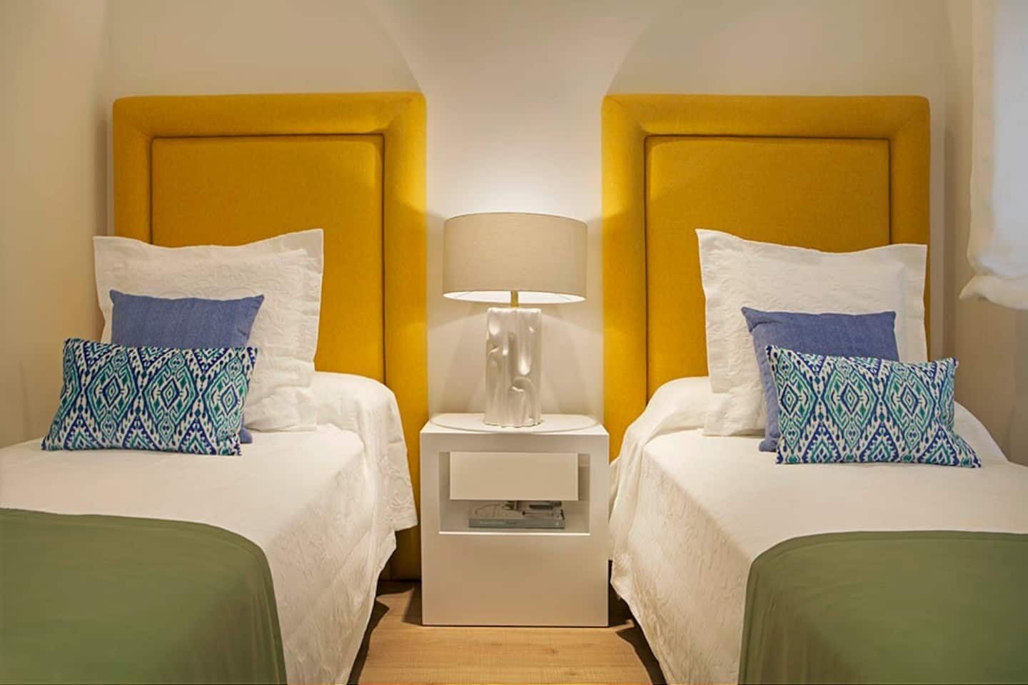 Habitación de hotel con dos camas, cada una con colcha blanca y almohadas estampadas en azul. Hay una mesita de noche con lámpara entre las camas.