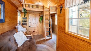 Interior de una cabaña de madera con una cama en altillo, un sofá, una escalera y una ventana con vistas.