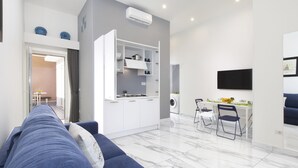 A modern living room with a blue sofa, a kitchen area with white cabinets, a dining table with chairs, and a flat-screen TV mounted on the wall.