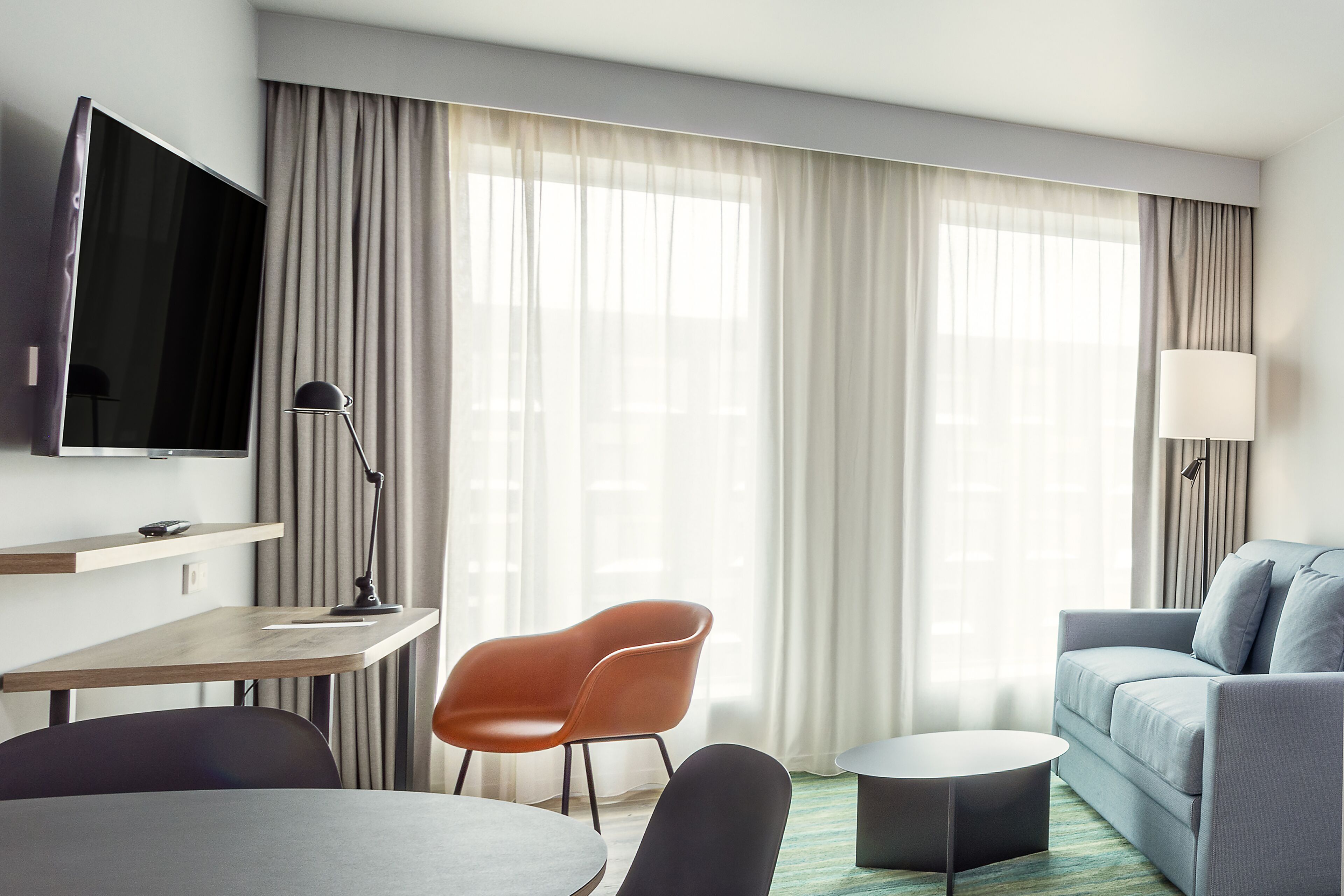 A modern hotel room with a flat-screen TV, a desk with a chair, a sofa, and a round coffee table.