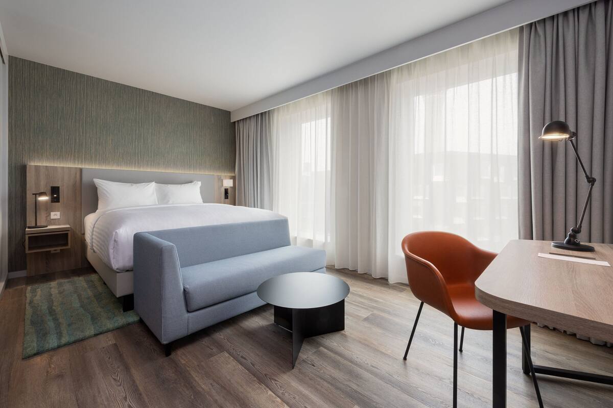 Ein modernes Hotelzimmer mit einem großen Bett, einem kleinen Sofa, einem runden Couchtisch, einem Esstisch mit einem Stuhl und einer Lampe.