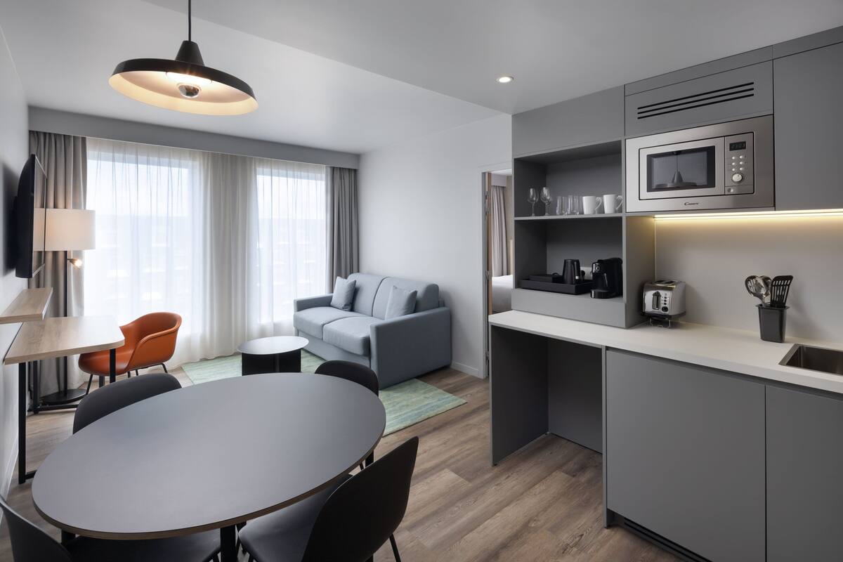 Ein modernes Hotelzimmer mit grauem Sofa, einem Esstisch und einer Küchenzeile inklusive Mikrowelle.