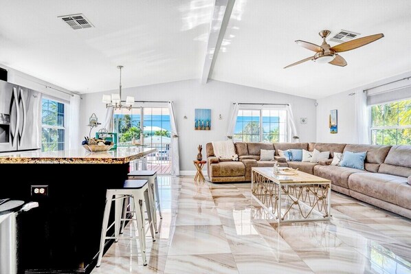 A spacious living area with a kitchen, a ceiling fan, and large windows offering a view of the outdoors.