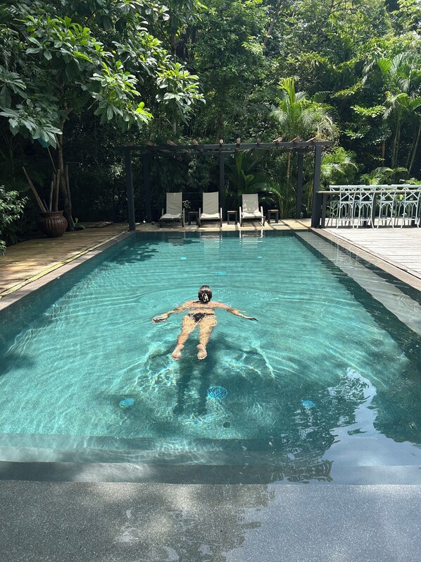 A clear swimming pool with a person floating, surrounded by lush greenery and shaded by trees.
