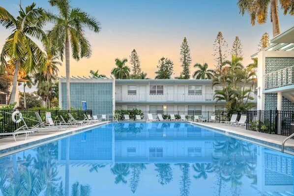 A swimming pool with lounge chairs and palm trees, surrounded by a building with a balcony and a fence. - Beautifully Updated Naples Condo, Walk to Beach & 3rd St (Naples)