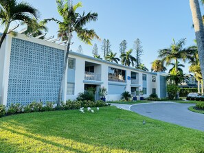 A modern building with a flat roof, multiple balconies, and a surrounding lawn with palm trees. - Beautifully Updated Naples Condo, Walk to Beach & 3rd St (Naples)
