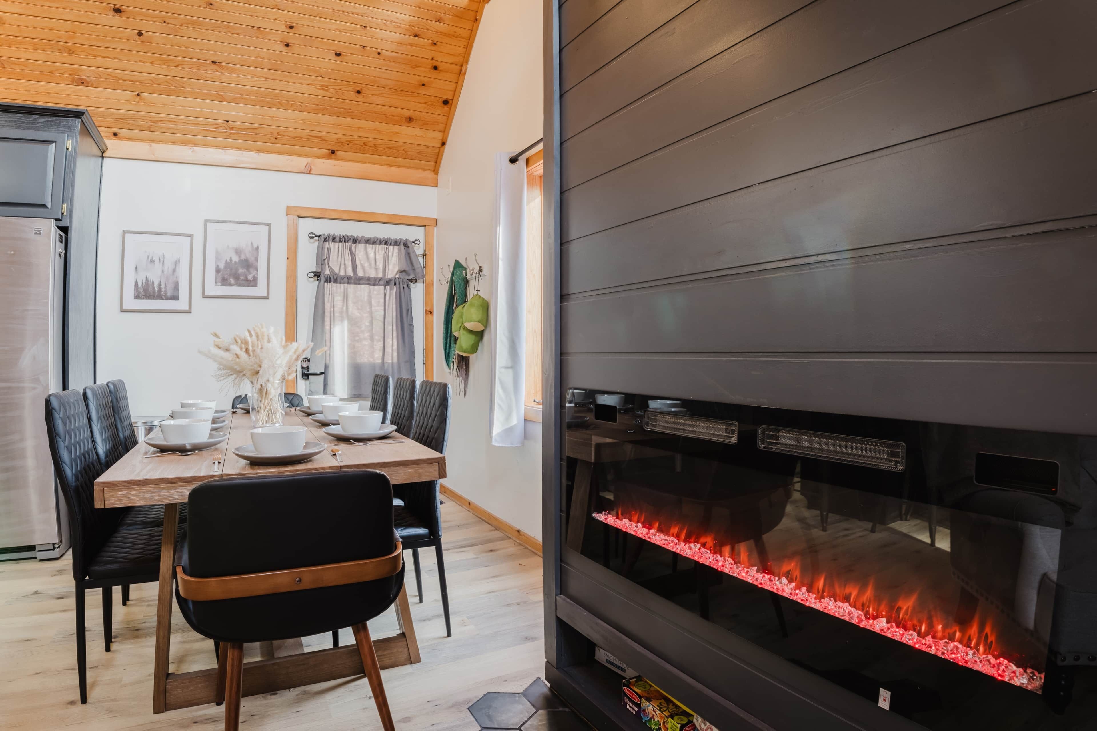 Un coin repas avec une table dressée pour quatre personnes, une cheminée moderne et une cuisine équipée d’un réfrigérateur.