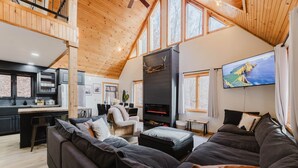 A spacious living area with a high ceiling, a fireplace, a large TV, and a kitchen in the background.