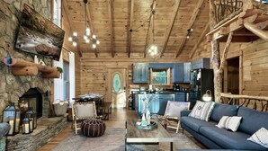 A rustic cabin interior with a stone fireplace, wooden ceiling beams, a dining area, and a living room with a sofa and coffee table. - Discounts 3+N 4BR Cabin Sleeps 10 Hot Tub, Game Room, Fire Pit, Fenced Yard-Pets (Blue Ridge)